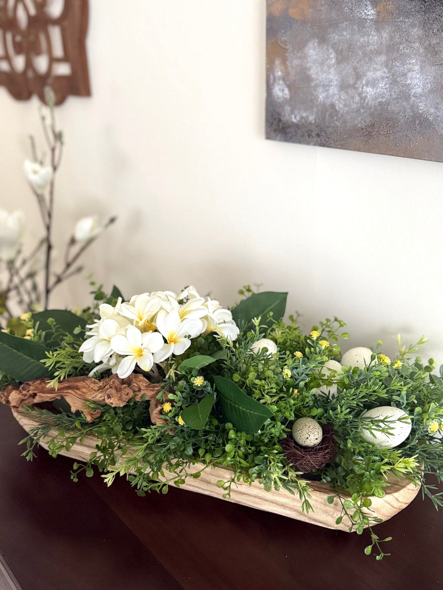 large easter centerpiece in earthy colors with white and yellow flowers, artificial greenery, faux ehhs, gfake nests and grapewood in long wooden decorative bowl