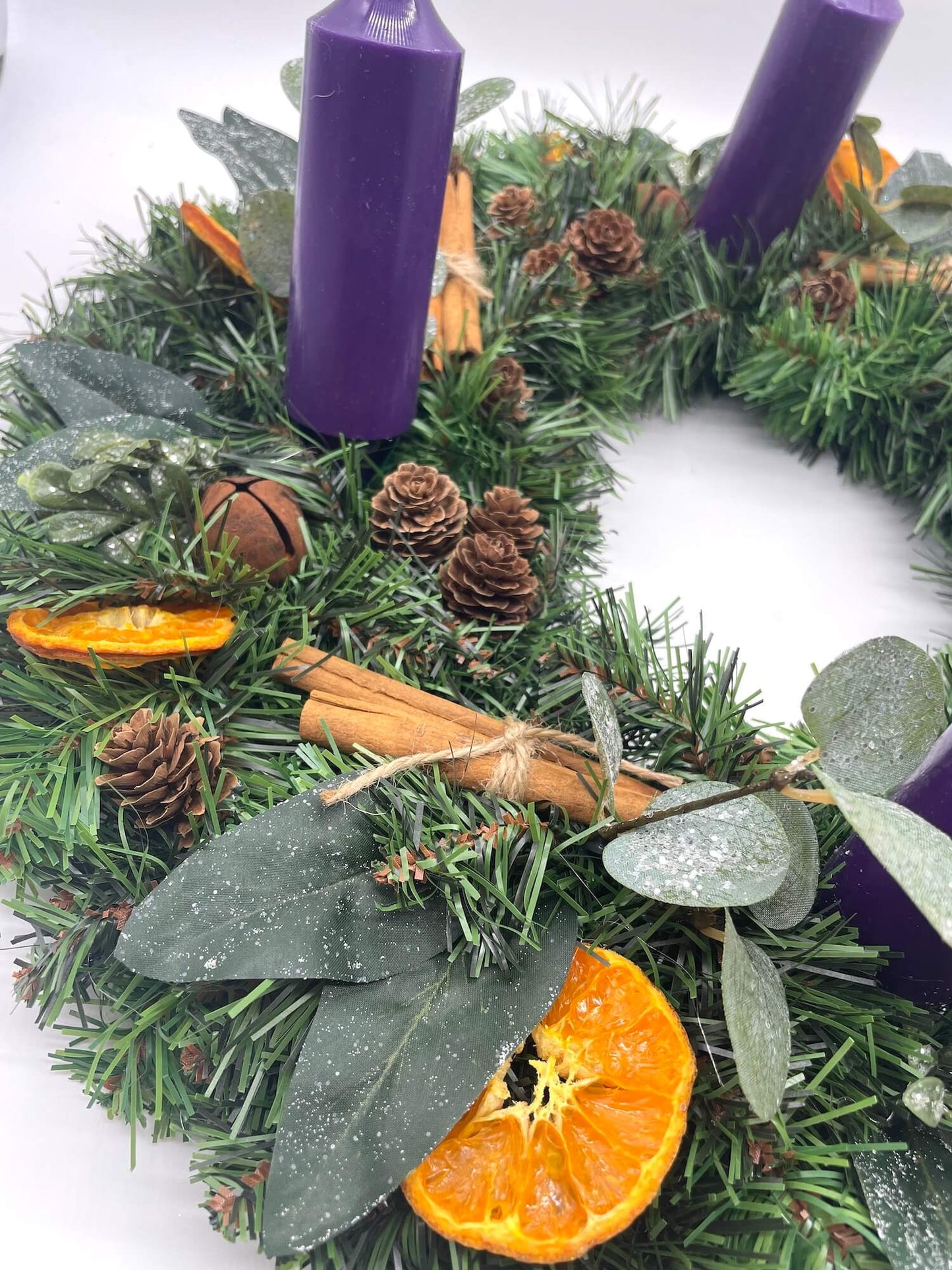 close up for Advent wreath with purple pink candles, pine cones, cinnamon sticks, and dried oranges, winter greenery, metal bells