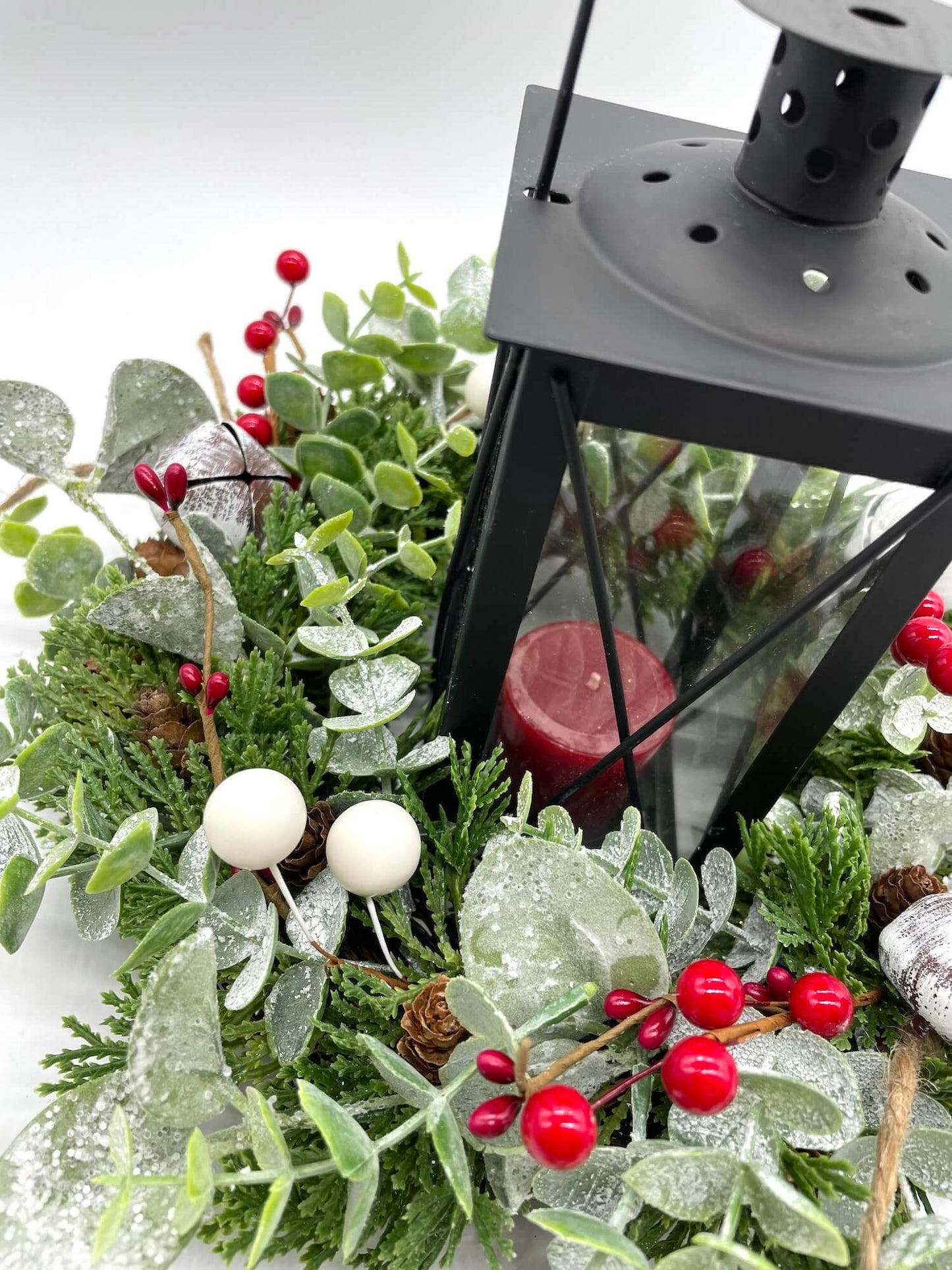 close up for winter evergreen table wreath with frosted greenery, white metal bells, red and white berries and black metal lantern in the middle