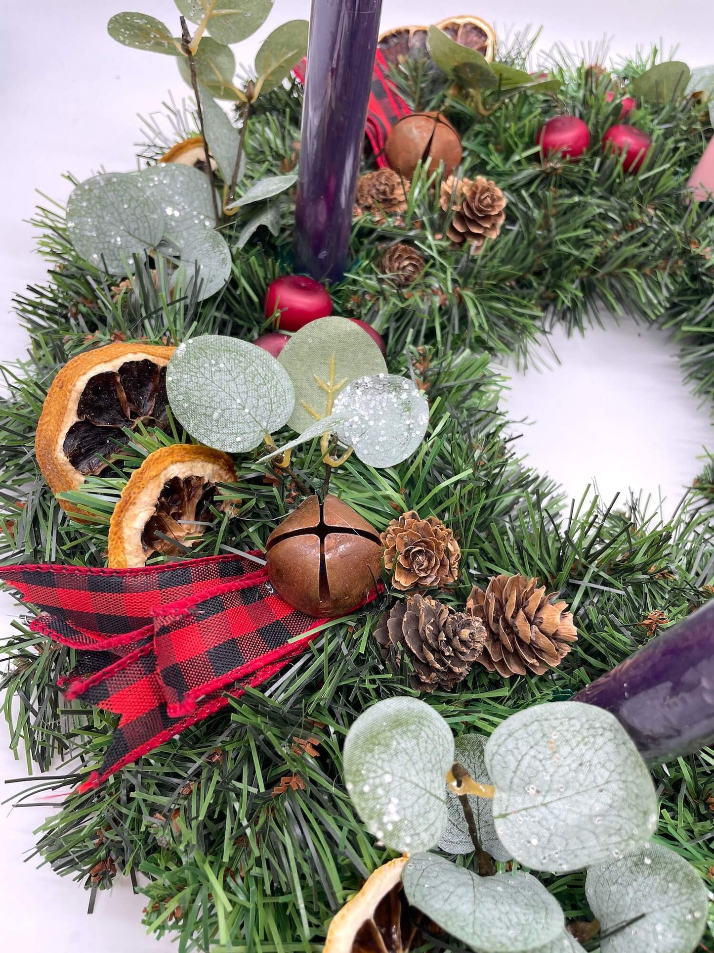 close up of Christmas wreath with pine cones, dried lemon slices, cranberries, metal bells, greenery and a red and black ribbon and purple pink tapper candles