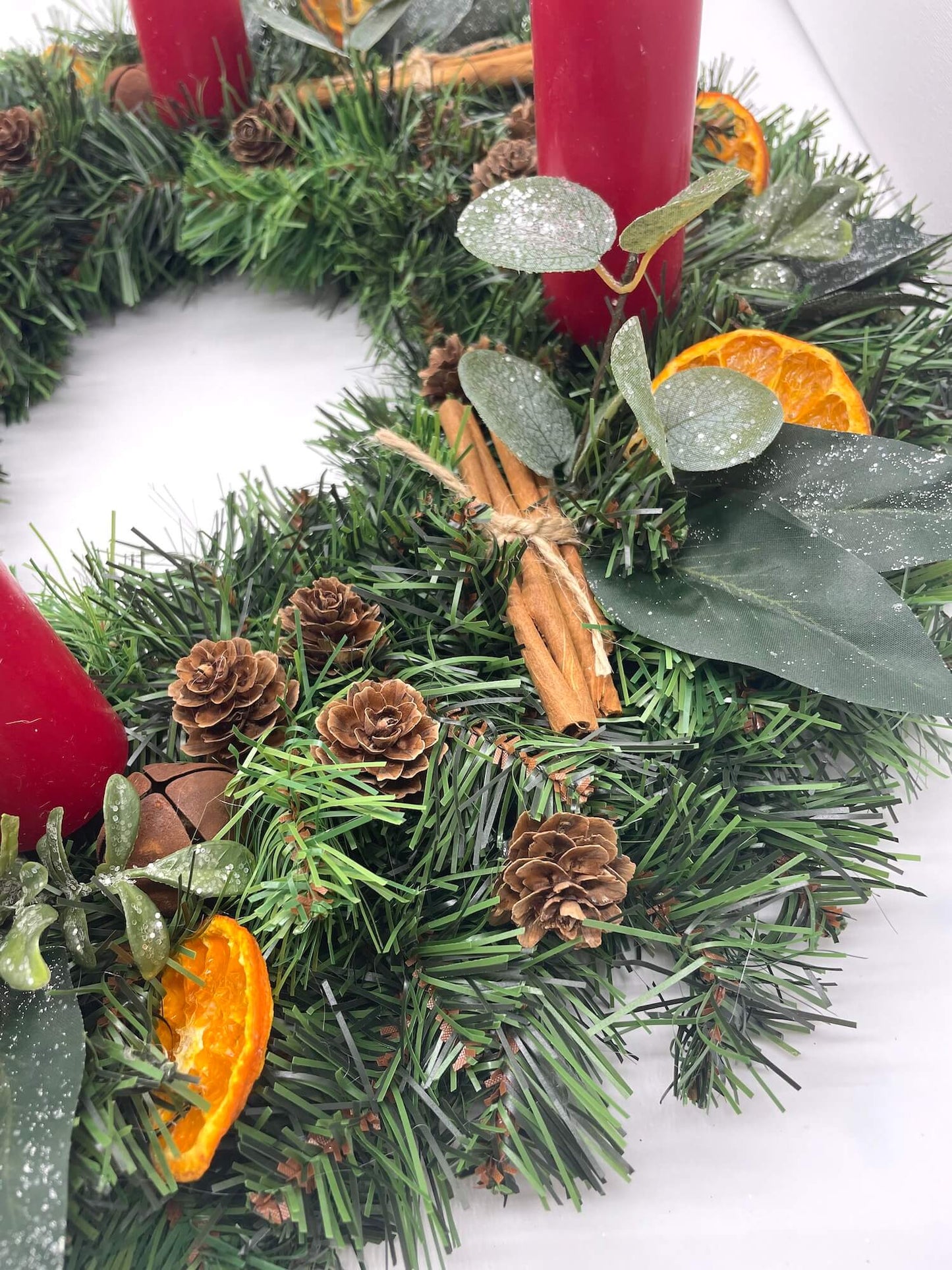 close up of advent wreath in rustic earthy style with cinnamon stick, dried orange slices, pinecones, winter greenery, metal bels and thick short red candles