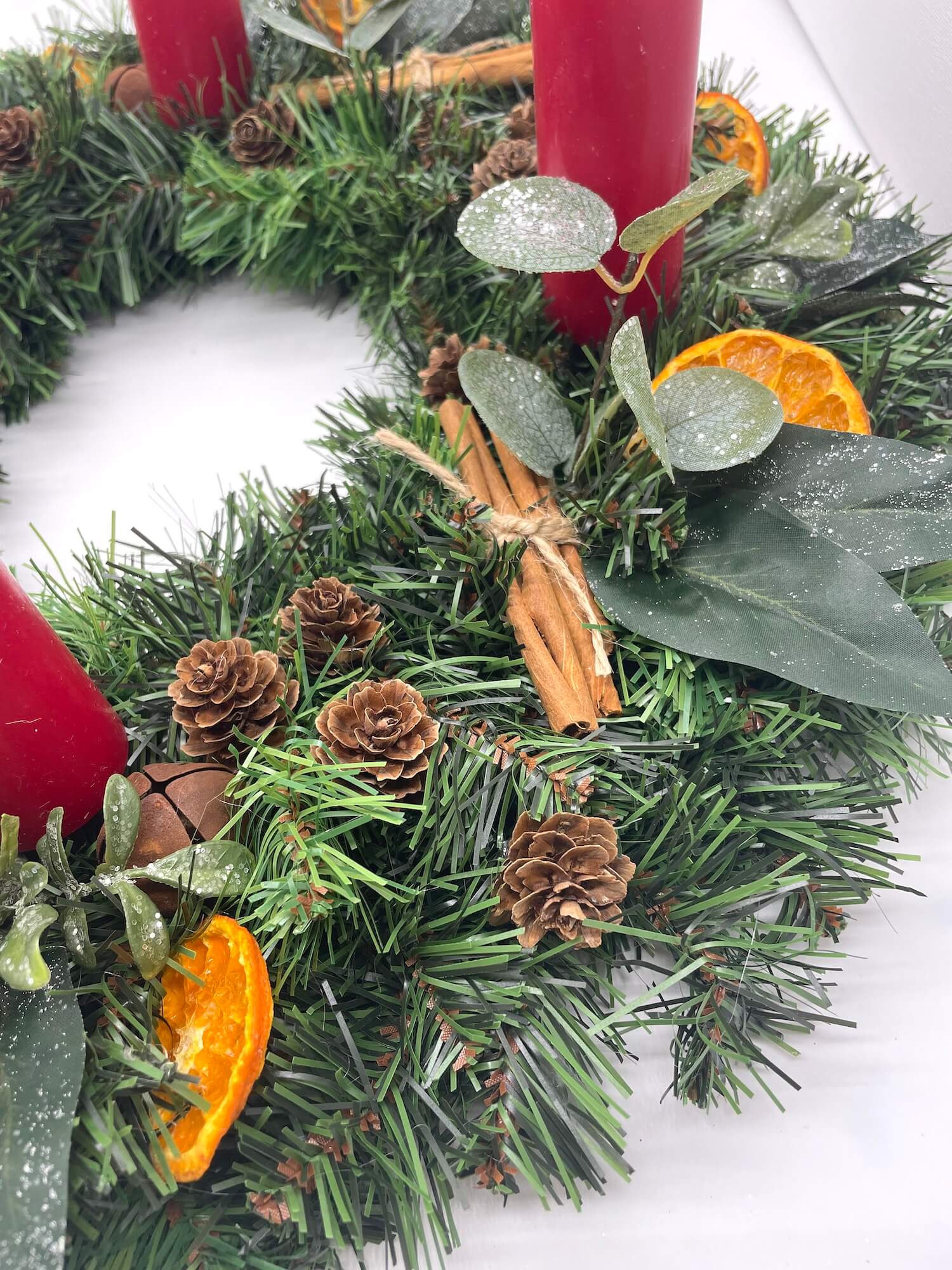 close up of advent wreath in rustic earthy style with cinnamon stick, dried orange slices, pinecones, winter greenery, metal bels and thick short red candles
