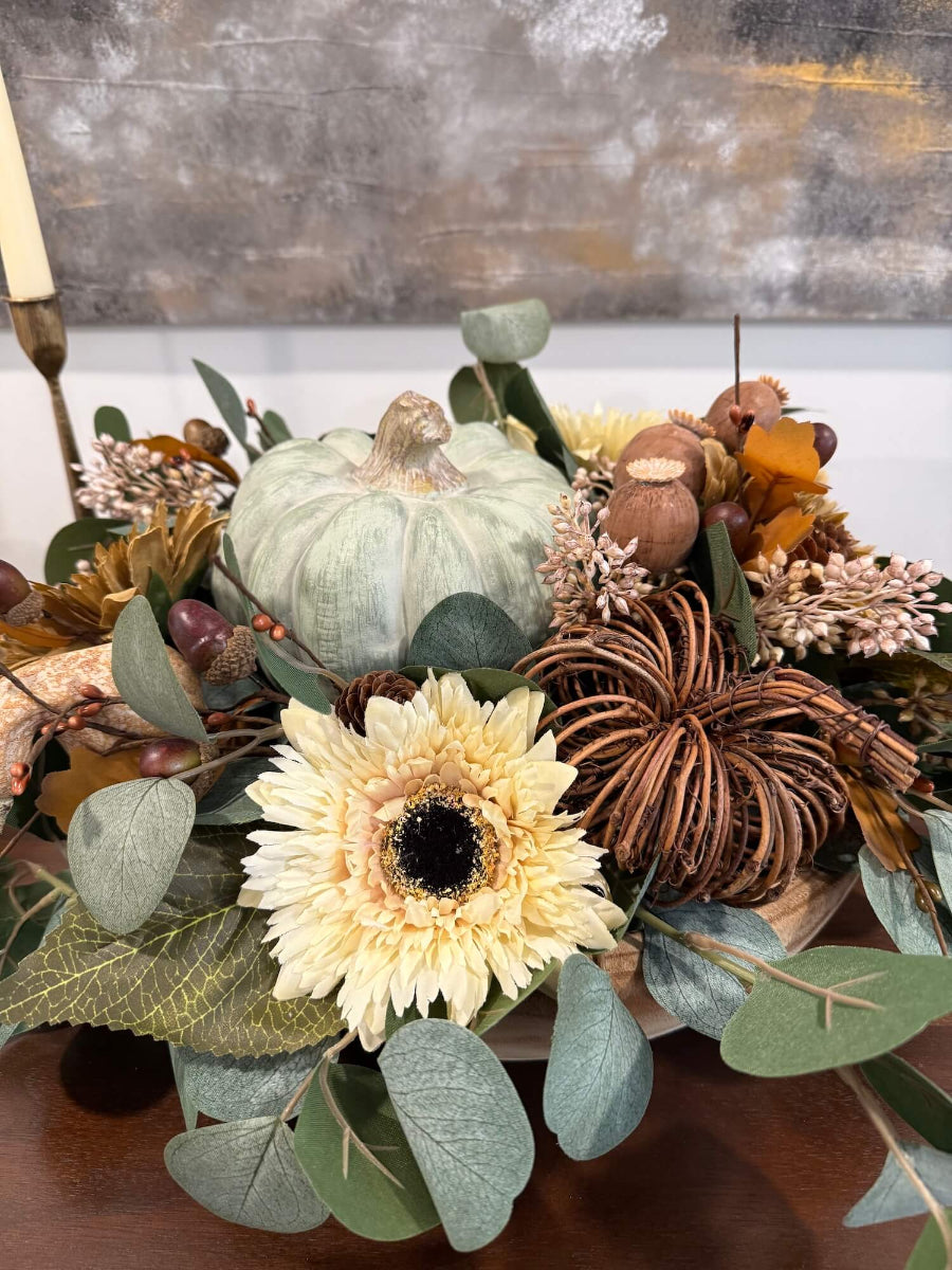 close view of elegant handmade fall centerpiece in neutral earthy colors with silver dollar eucalyptus stems, fall flowers in cream and beige/brown, grapevine pumpkin, cream/beige small pumpkin, wheat, poppy heads and acorn stems with wooden dusty green pumpkin in the middle, all set in round wooden vessel