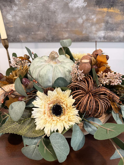 close view of elegant handmade fall centerpiece in neutral earthy colors with silver dollar eucalyptus stems, fall flowers in cream and beige/brown, grapevine pumpkin, cream/beige small pumpkin, wheat, poppy heads and acorn stems with wooden dusty green pumpkin in the middle, all set in round wooden vessel