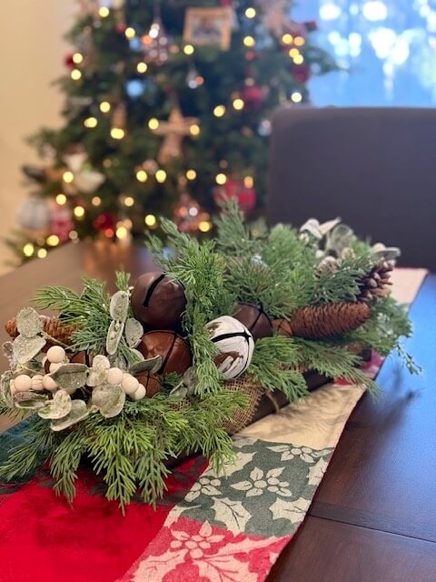 Decorative Christmas centerpiece with variety of greenery, pinecones, and jingle bells in long wooden planter