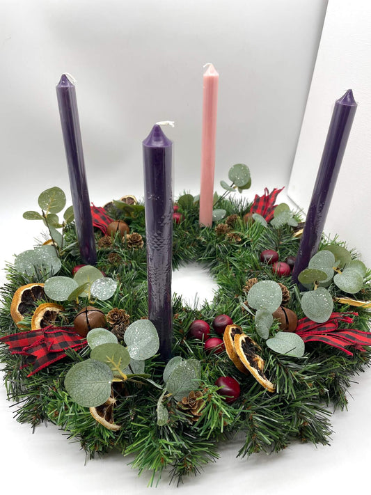 evergreen Advent wreath with purple pink candles decorated with greenery, dried lemons, cranberry, metal bels and pinecones, red-black ribbon