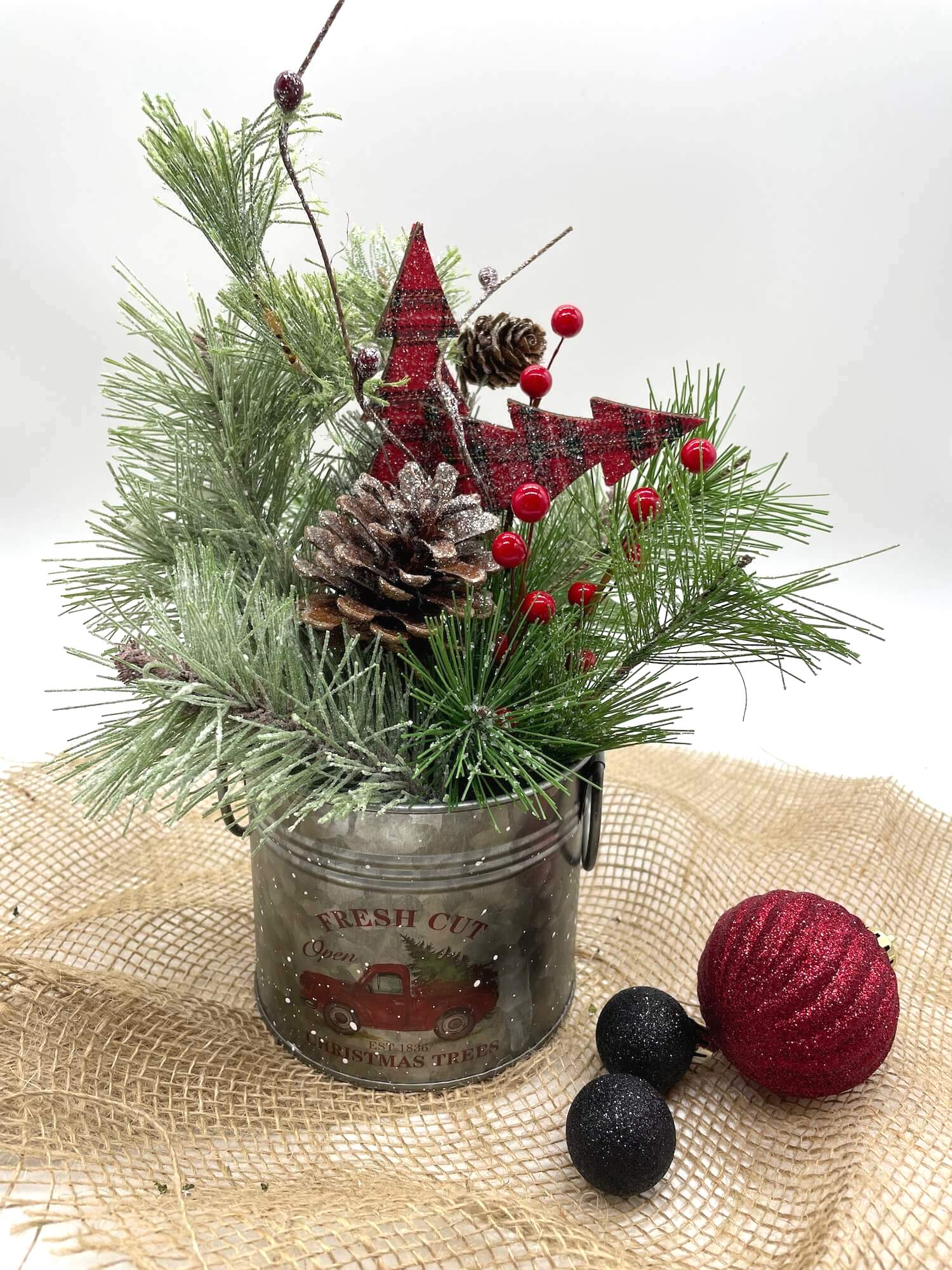  Christmas arrangement with variety of winter greenery, red ornaments, pinecones in a metal tin with red truck