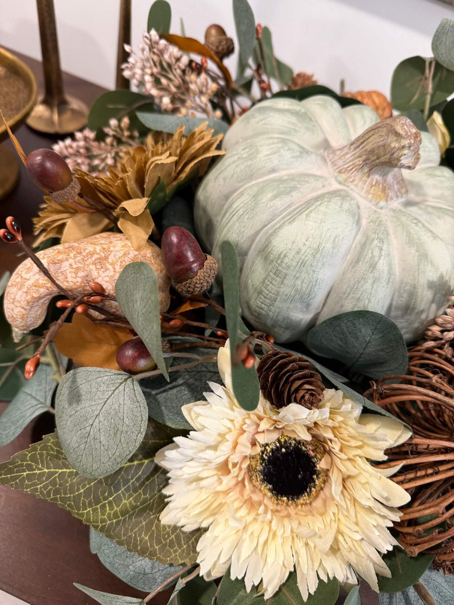 close up for elegant handmade fall centerpiece in neutral earthy colors with silver dollar eucalyptus stems, fall flowers in cream and beige/brown, grapevine pumpkin, cream/beige small pumpkin, wheat, poppy heads and acorn stems with wooden dusty green pumpkin in the middle, all set in round wooden vessel