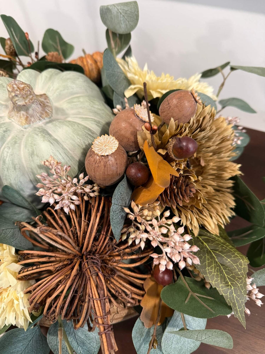 close up for elegant handmade fall centerpiece in neutral earthy colors with silver dollar eucalyptus stems, fall flowers in cream and beige/brown, grapevine pumpkin, cream/beige small pumpkin, wheat, poppy heads and acorn stems with wooden dusty green pumpkin in the middle, all set in round wooden vessel