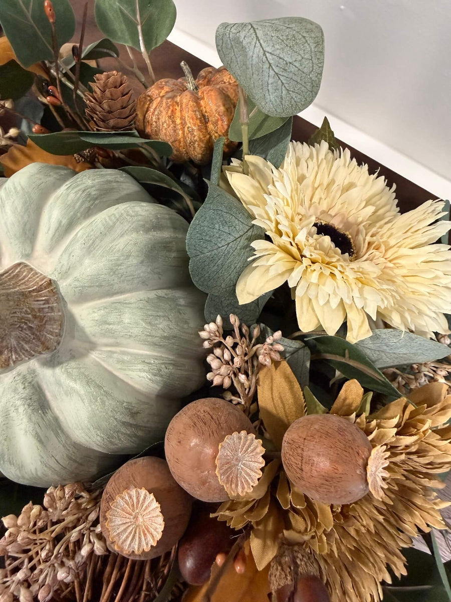close up for elegant handmade fall centerpiece in neutral earthy colors with silver dollar eucalyptus stems, fall flowers in cream and beige/brown, grapevine pumpkin, cream/beige small pumpkin, wheat, poppy heads and acorn stems with wooden dusty green pumpkin in the middle, all set in round wooden vessel