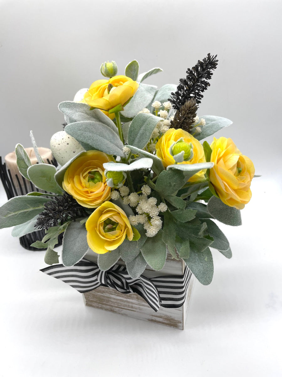 Easter Arrangement handmade with yellow flower accompanied by greenery, white flower and completed with faux eggs placed in whitewashed wooden vase decorated with balc white striped ribbon