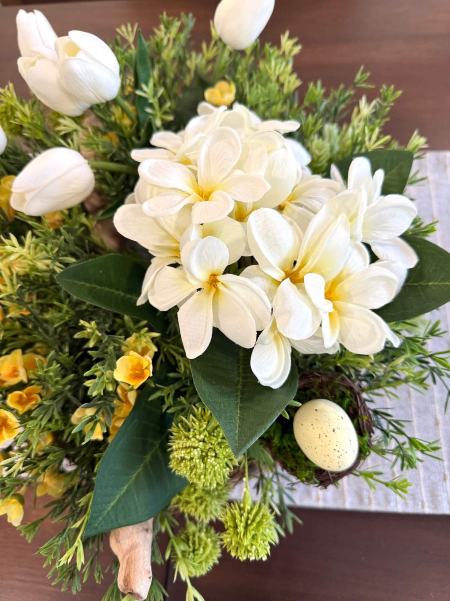 close up for white primrose, white tulips and yellow flowers, greenery and faux egg in nest in easter spring centerpiece