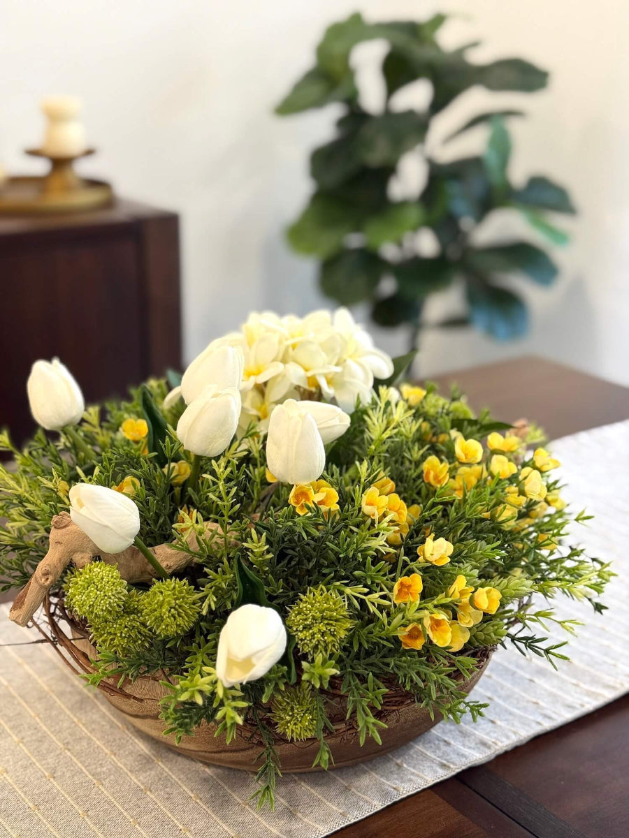 lush elegant spring easter centerpiece with vivid greenery, white tulips, white primrose, yellow flowers and driftwood in round wooden dough now decorated with grapevine