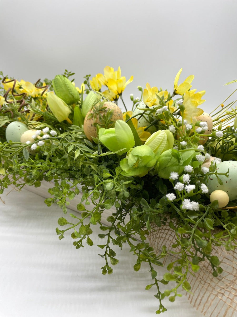 easter centerpiece with faux green and yellow tulips, realistic greenery and white flowers, faux eggs in whitewashe wooden planter decorated with burlap