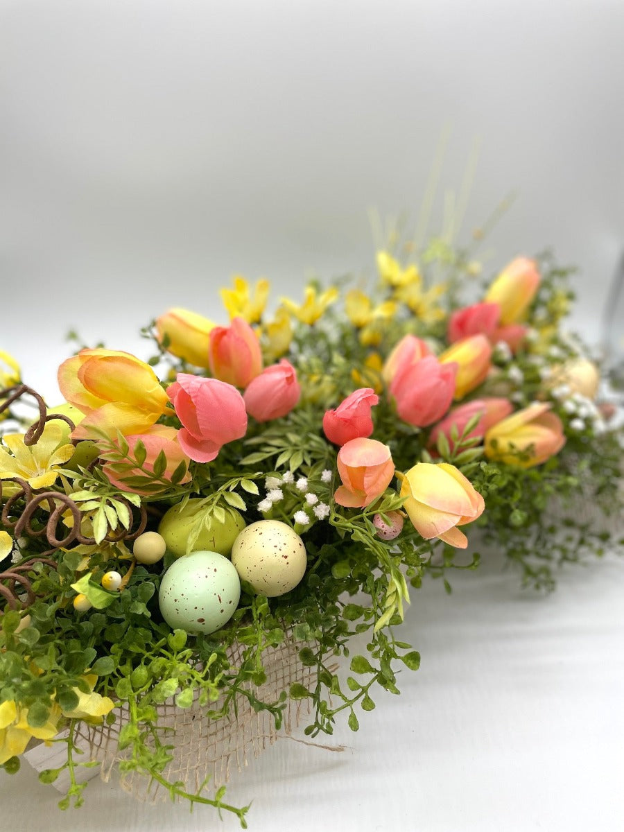 Easter Centerpiece  with pink and yello tulips, variety of greenery, delicate white flowers, faux eggs in whitewashed wooden planter decorated with burlap
