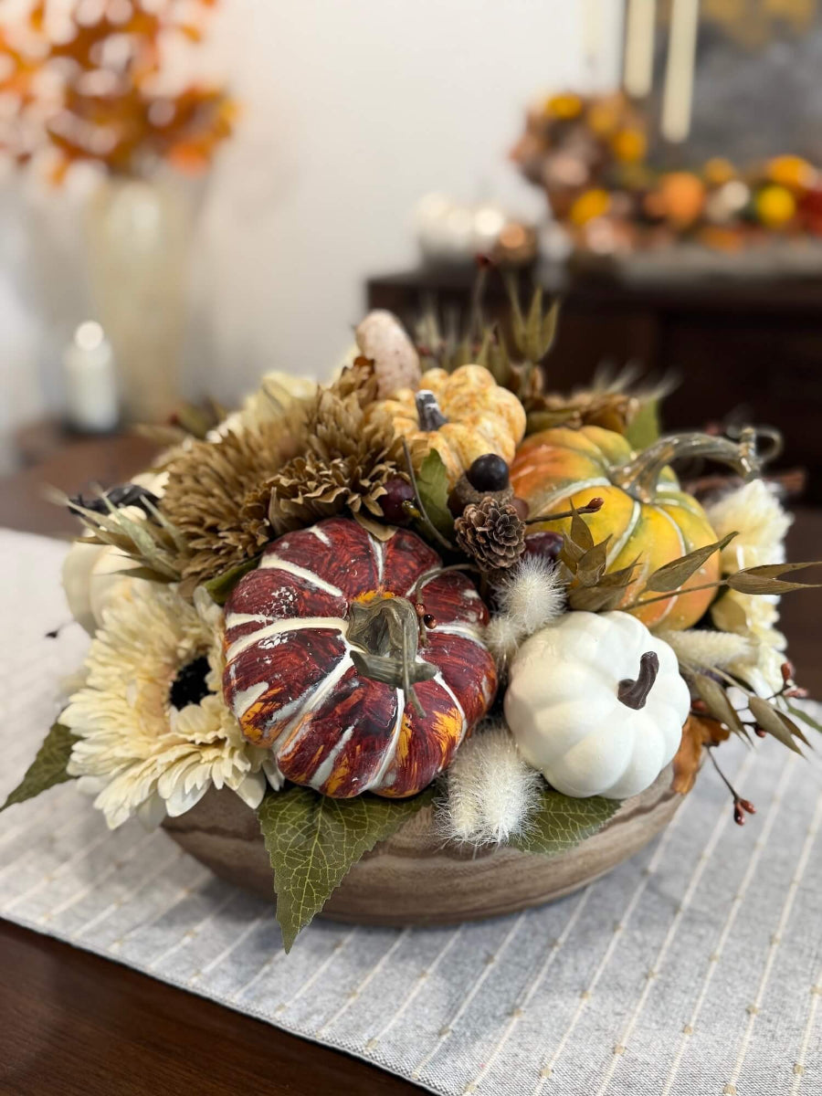 round fall centerpiece in neutral colors with faux pumpkins, wheat stems, fall flowers and leaves, and acorn stems in round wooden vessel set on dining table