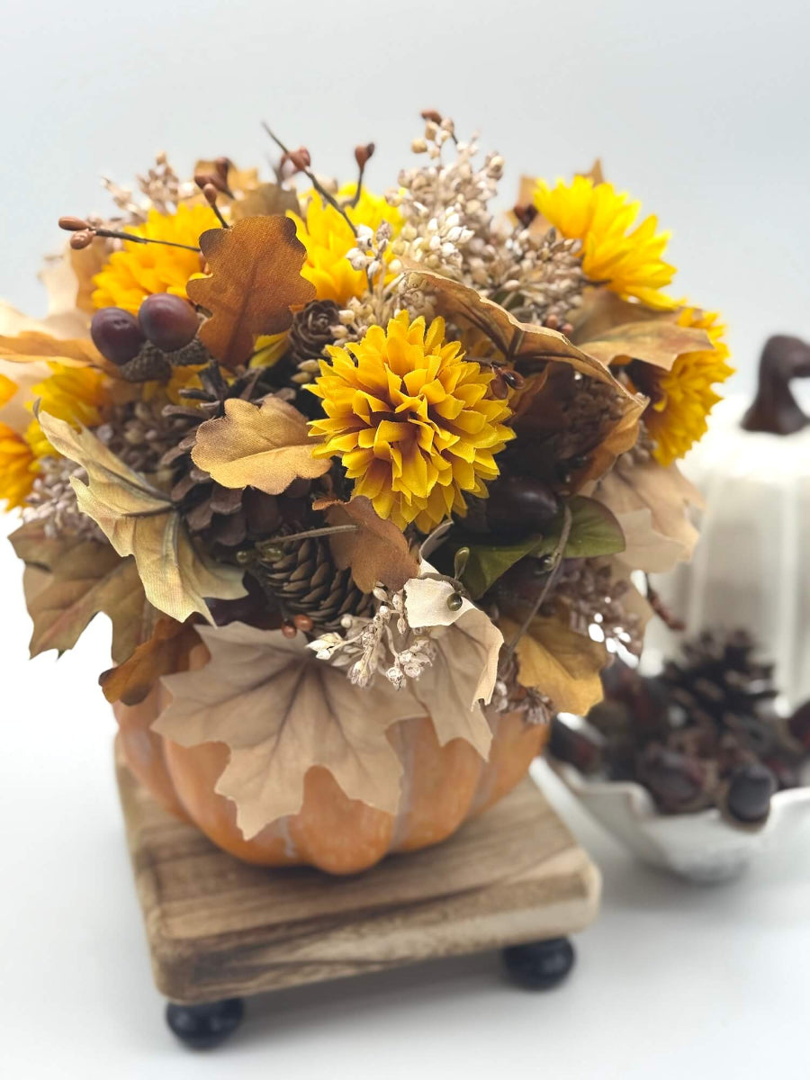 autumn arrangement with flowers, leaves, pinecones, wheat stems in ywllow, beige, brown colors on dusty orange wooden pumpkin