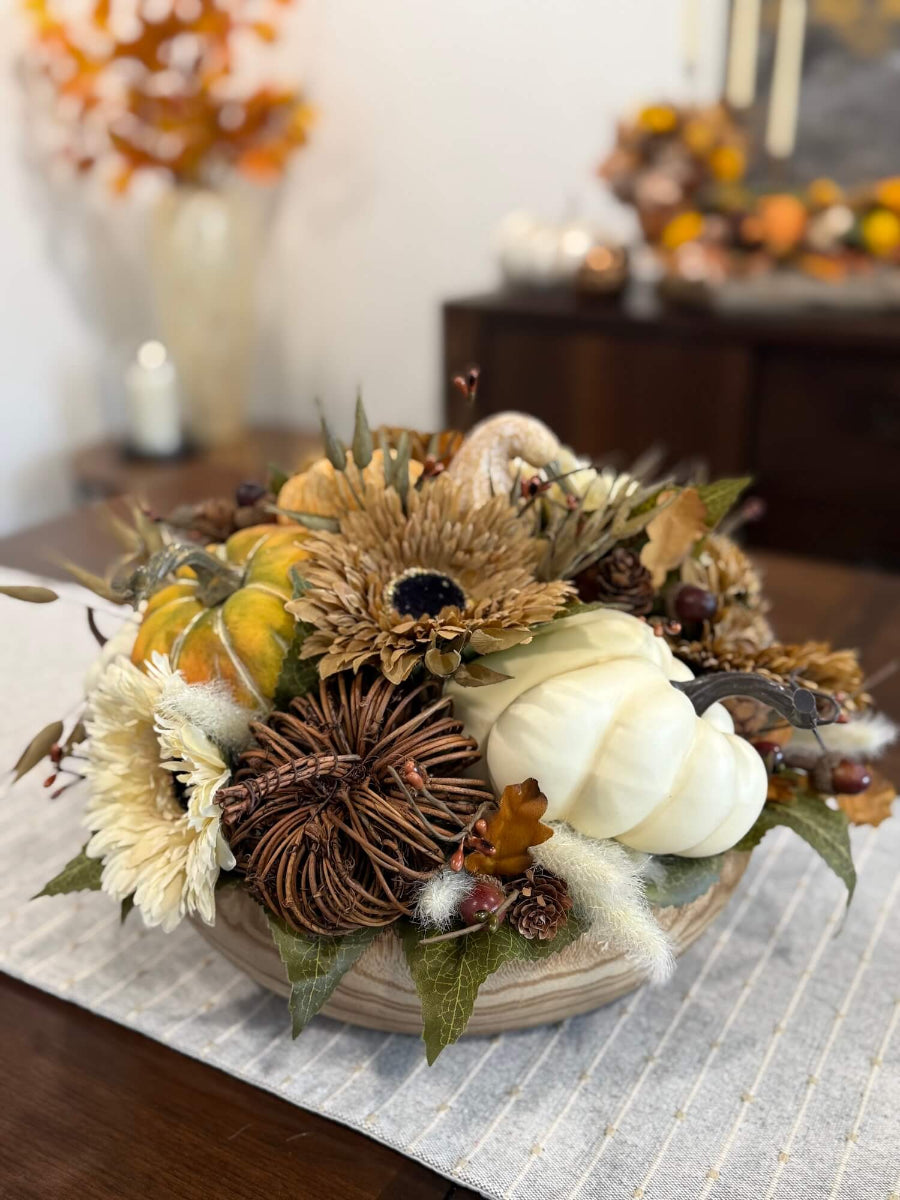 round fall centerpiece in neutral colors with faux pumpkins, wheat stems, fall flowers and leaves, and acorn stems in round wooden vessel set on dining table