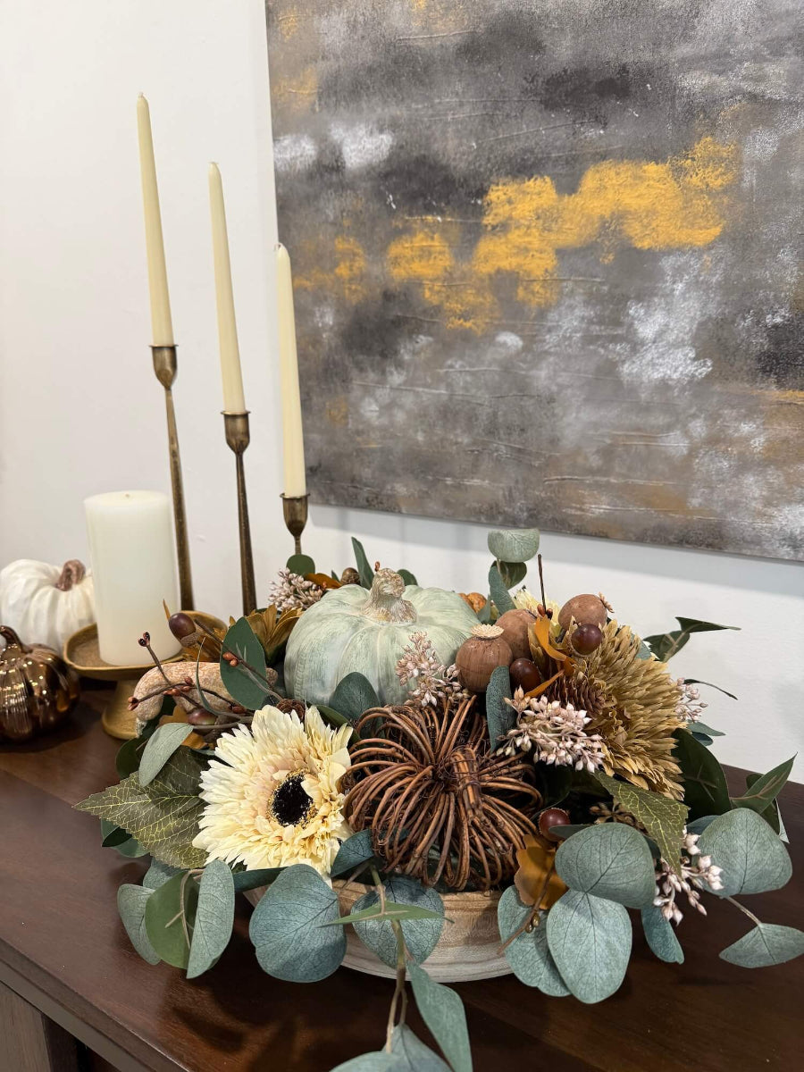 elegant handmade fall centerpiece in neutral earthy colors with silver dollar eucalyptus stems, fall flowers in cream and beige/brown, grapevine pumpkin, cream/beige small pumpkin, wheat, poppy heads and acorn stems with wooden dusty green pumpkin in the middle, all set in round wooden vessel