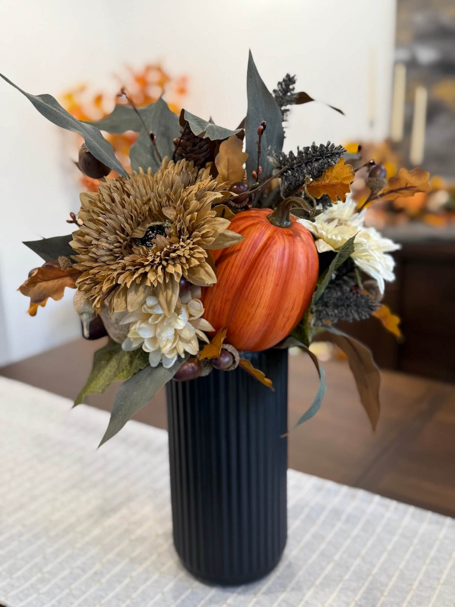 tall fall centerpiece with fall flowers, leaves, black what stems, poppy heads, acorn stems and pumpkins in black textured ceramic tall vase set on a table