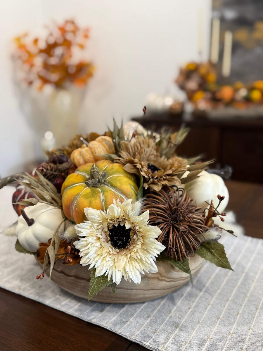 round fall centerpiece in neutral colors with faux pumpkins, wheat stems, fall flowers and leaves, and acorn stems in round wooden vessel set on dining table
