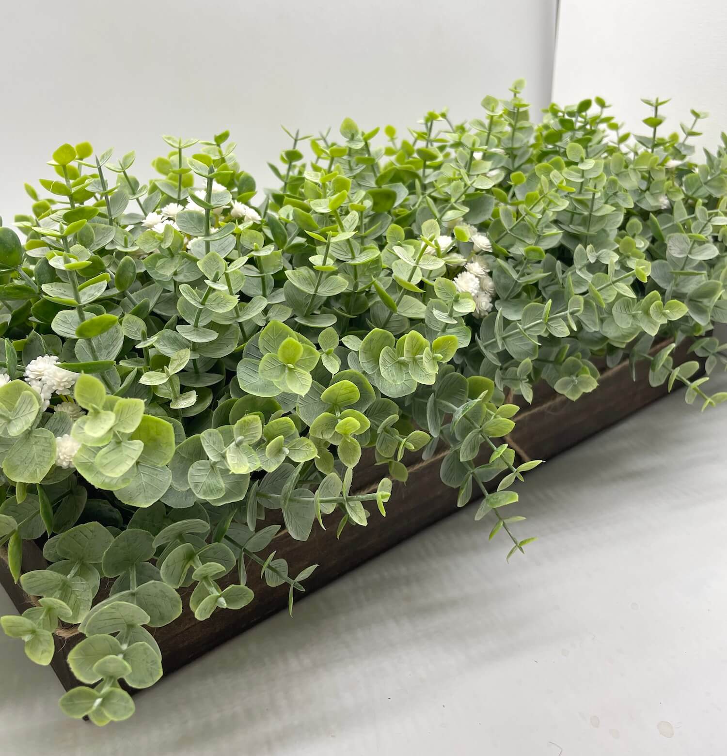realistic looking faux eucalyptus arrangement with delicate white flowers in long wooden planter stained in espresso color and decorated with jute