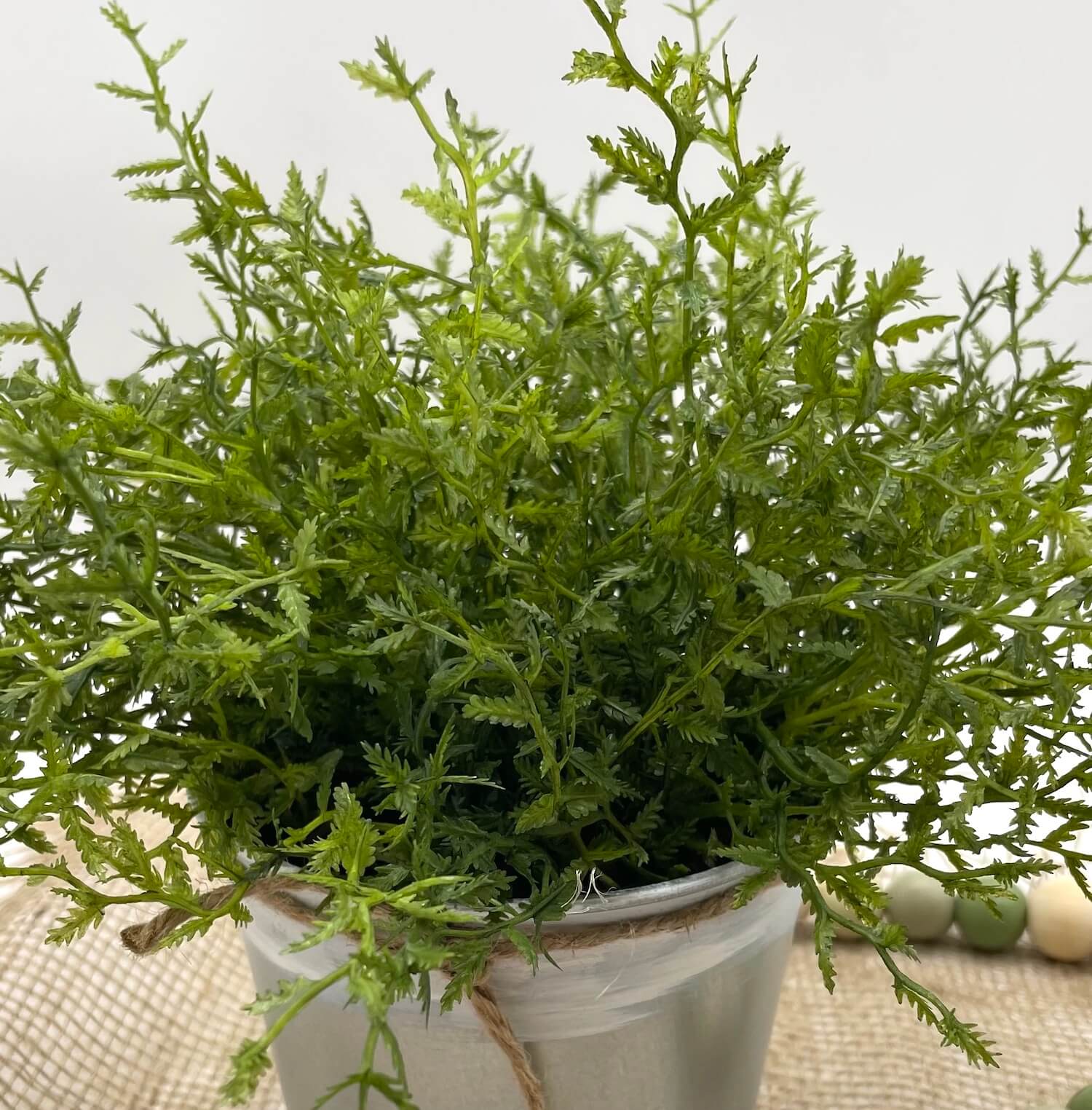 close up vivid green faux plant fern in metal pot touched with white paint and decorated with jute