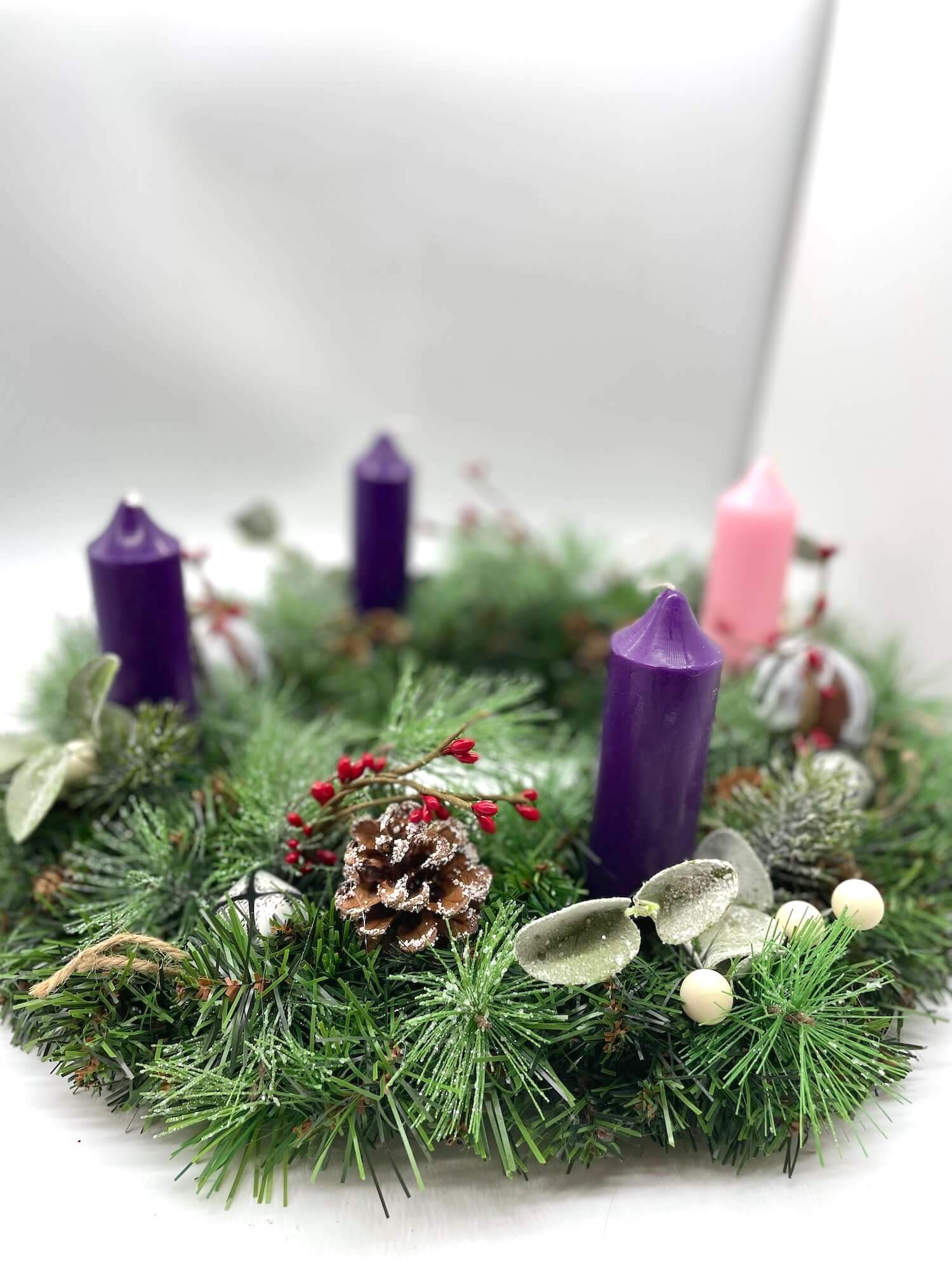 handcrafted advent wreath made of evergreen, variety of winter greenery, pinecones, red and white berries and metal bells with short thick purple and pink candles
