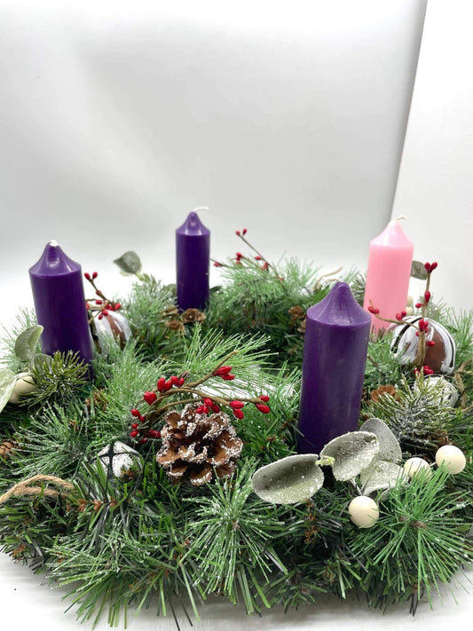 handcrafted advent wreath created out of evergreen, variety of winter greenery, pinecones, red and white berries and metal bells with short thick purple and pink candles