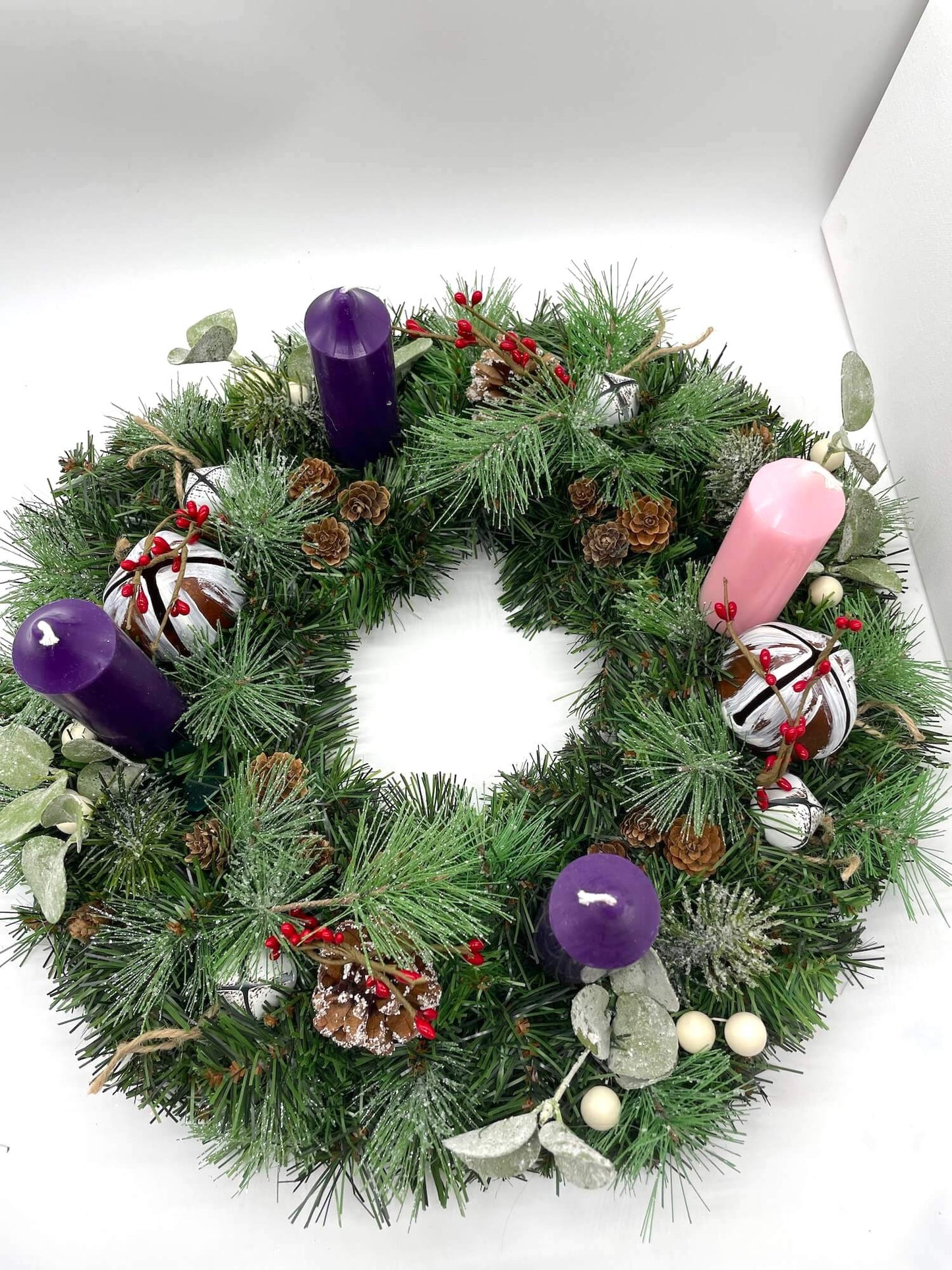 view from top of handcrafted advent wreath created out of evergreen, variety of winter greenery, pinecones, red and white berries and metal bells with short thick purple and pink candles