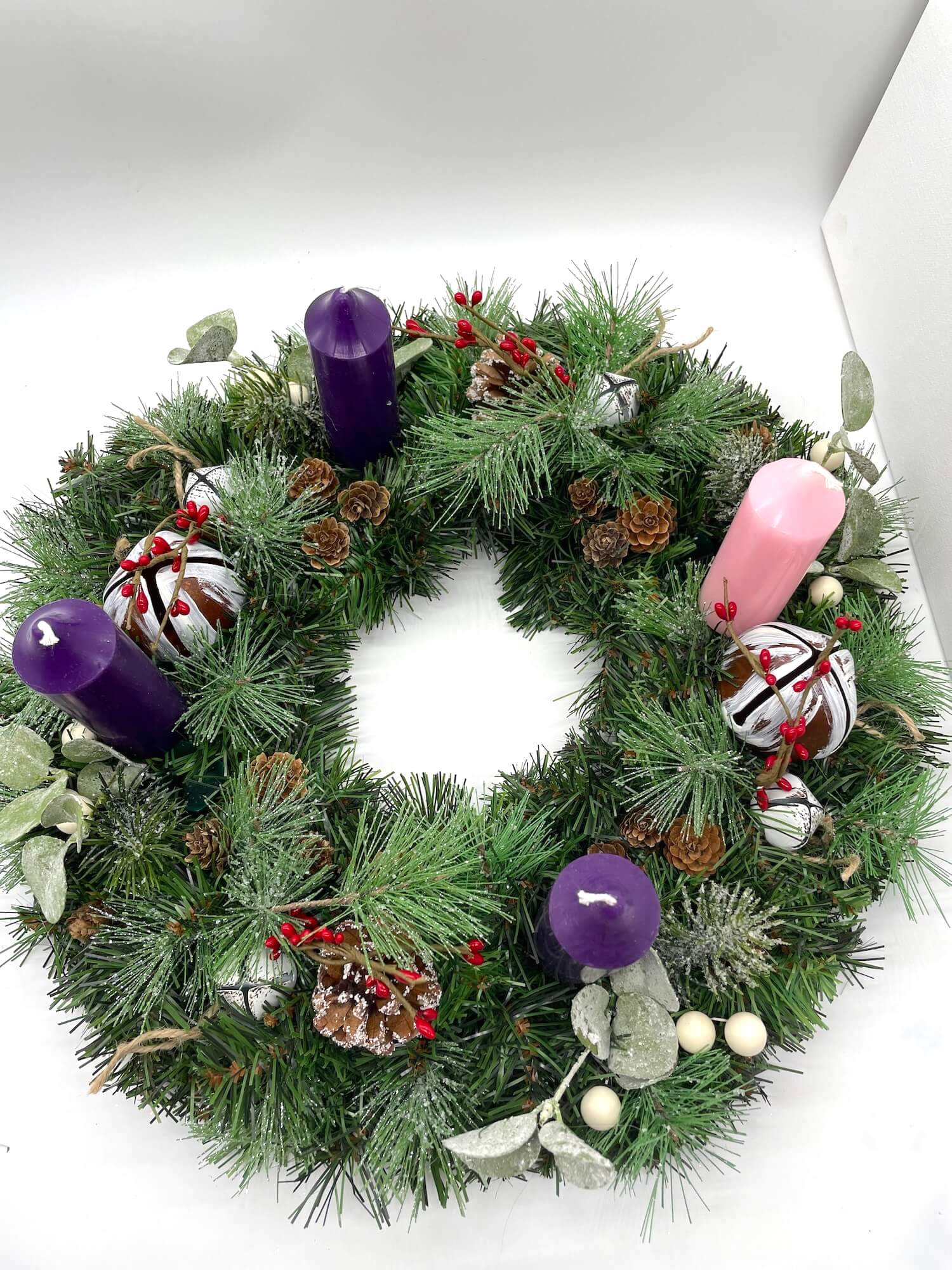 view from top of handcrafted advent wreath created out of evergreen, variety of winter greenery, pinecones, red and white berries and metal bells with short thick purple and pink candles