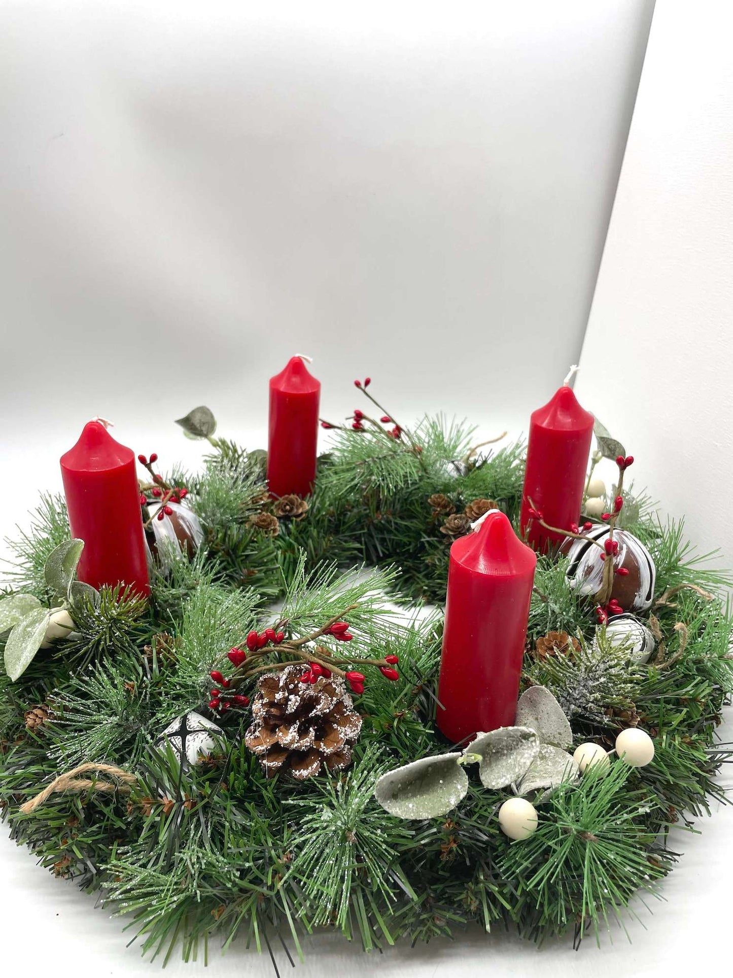 handcrafted advent wreath with evergreen, variety of winter greenery, pinecones, red and white berries and metal bells with short thick red candles