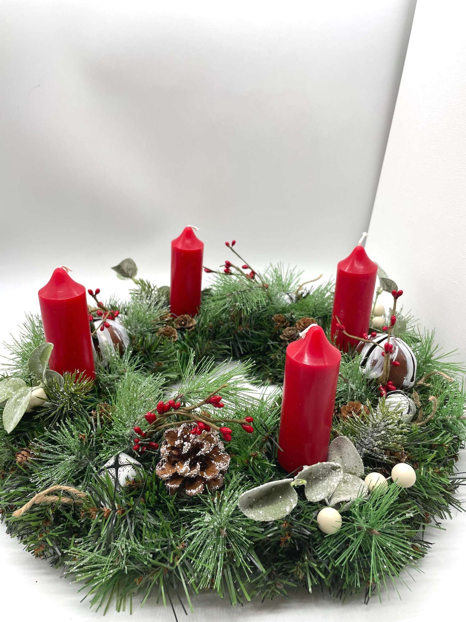 handcrafted advent wreath with evergreen, variety of winter greenery, pinecones, red and white berries and metal bells with short thick red candles