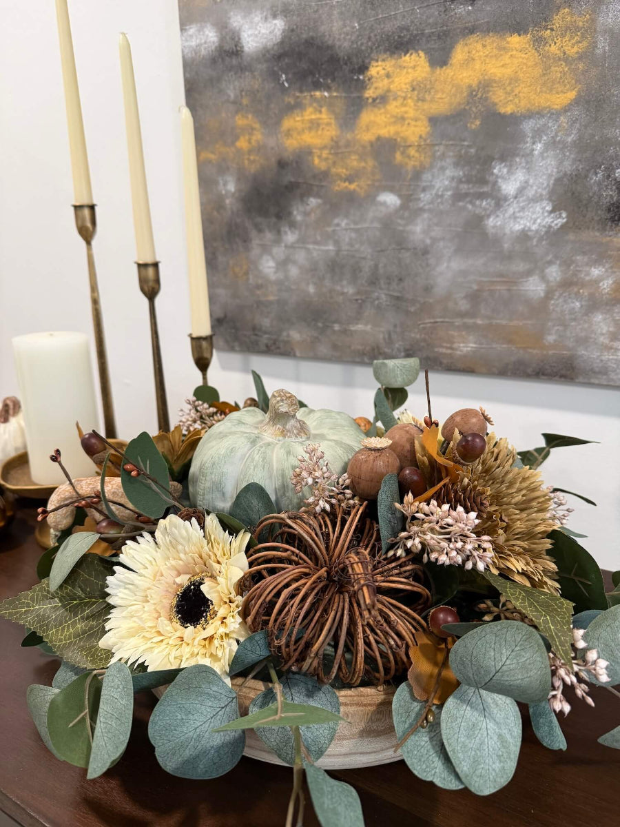 elegant handmade fall centerpiece in neutral earthy colors with silver dollar eucalyptus stems, fall flowers in cream and beige/brown, grapevine pumpkin, cream/beige small pumpkin, wheat, poppy heads and acorn stems with wooden dusty green pumpkin in the middle, all set in round wooden vessel