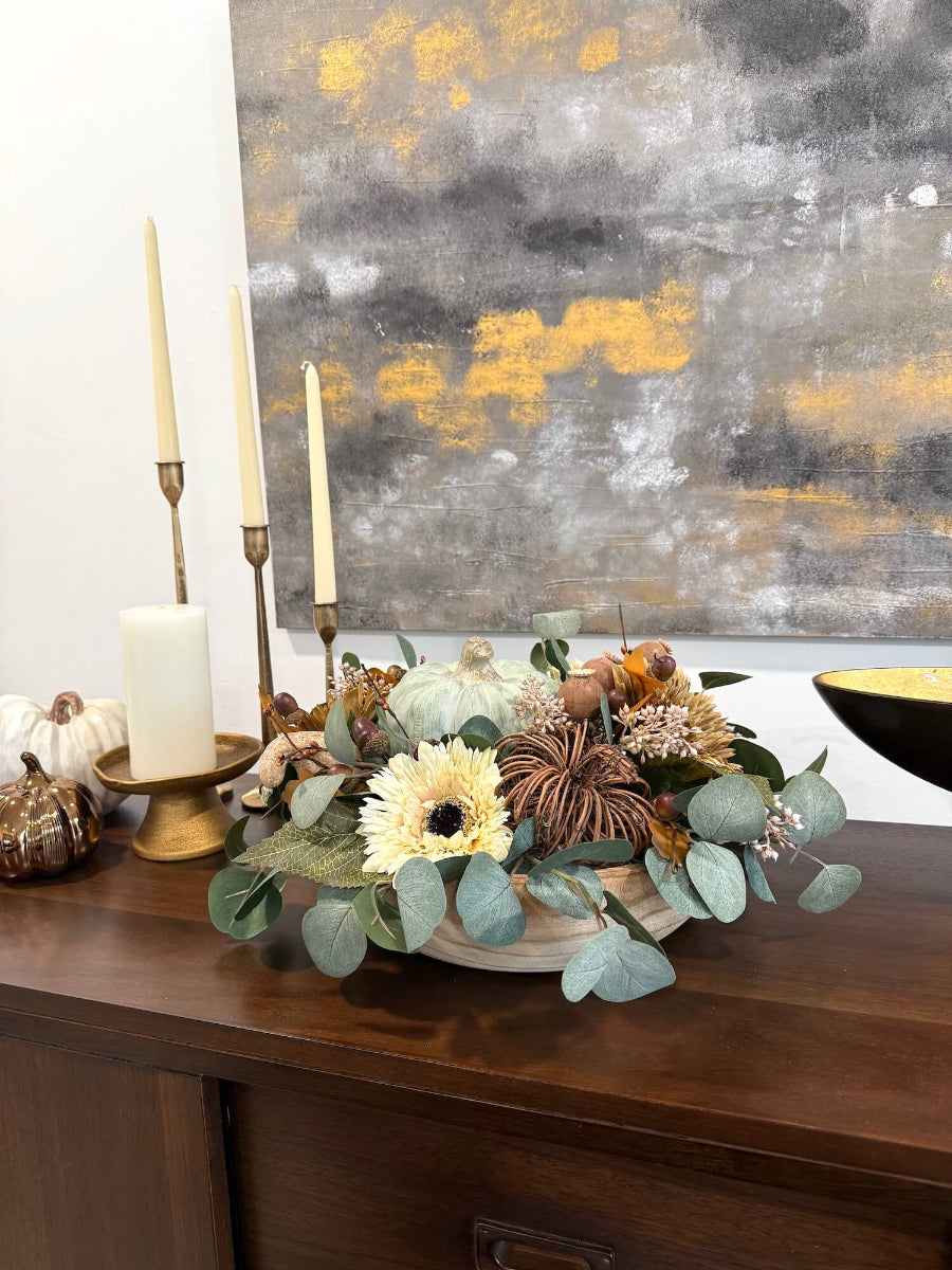 view on console table of elegant handmade fall centerpiece in neutral earthy colors with silver dollar eucalyptus stems, fall flowers in cream and beige/brown, grapevine pumpkin, cream/beige small pumpkin, wheat, poppy heads and acorn stems with wooden dusty green pumpkin in the middle, all set in round wooden vessel