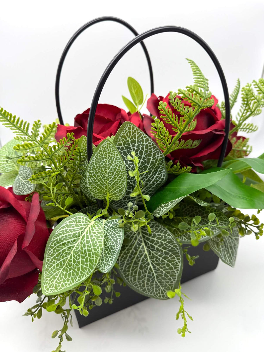 Floral arrangement with red roses and variety of greenery in a black gift bag
