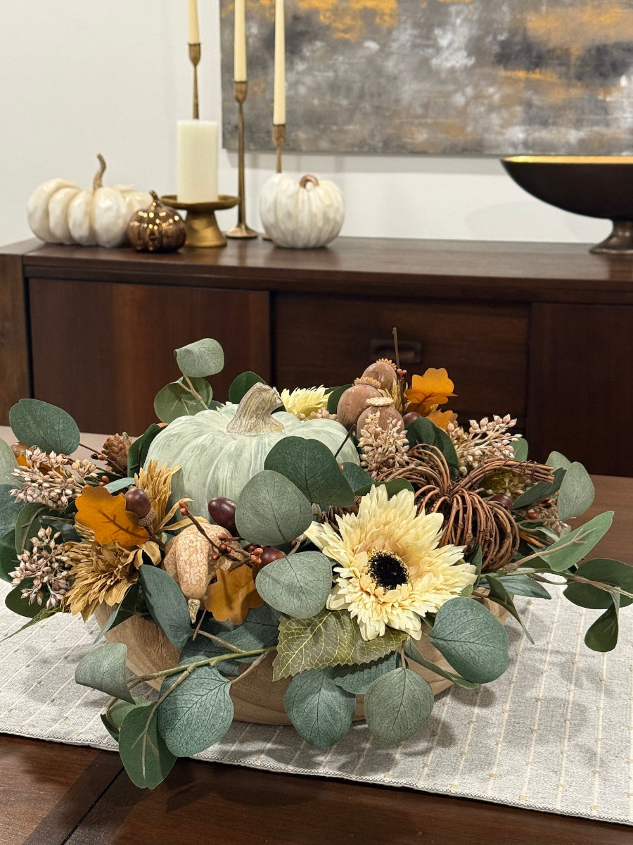 elegant handmade round fall centerpiece in neutral earthy colors with dusty green wooden pumpkin in middle, fall flowers in beige and cream,  silver dollar eucalyptus stems, wheat and pumpkins in beige, grapevine pumpkin and acorns, all in round wooden vessel 