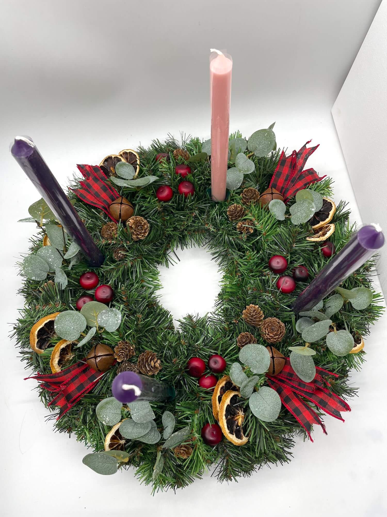 top view of advent wreath with natural elements of cranberries, dried lemon slices, pinecones, metal bells and red-black ribbon for rustic look with purple pink tapper candles