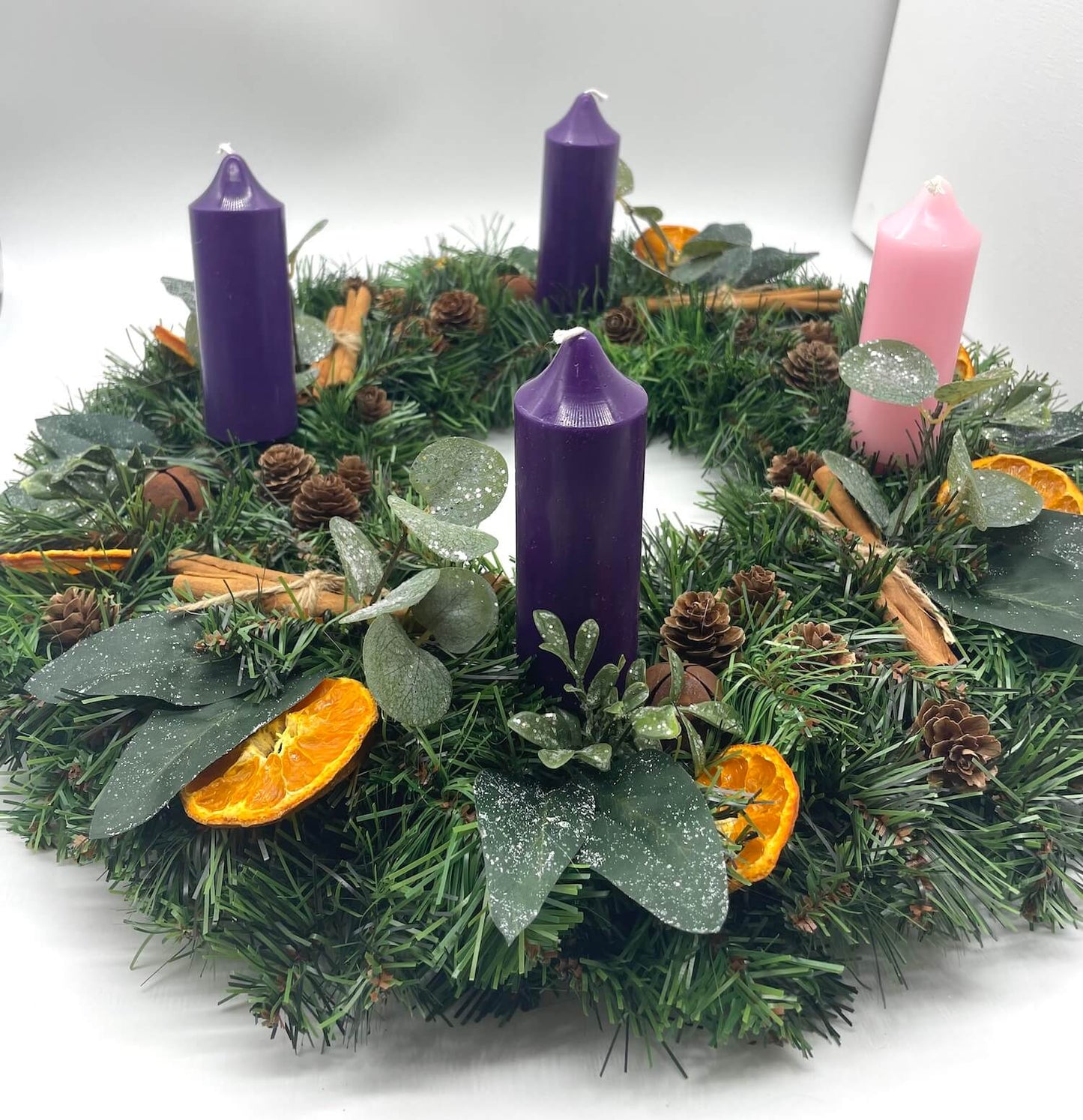 rustic Advent wreath with pink purple candles and earthy elements of orange dried slices, cinnamon sticks, pinecones, brown metal bells and winter greenery