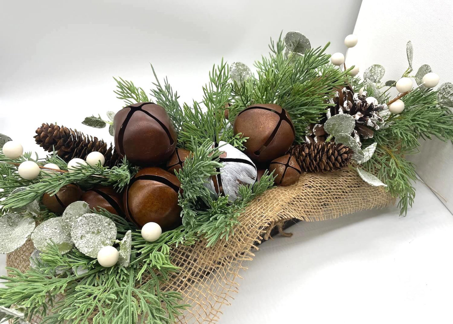 winter centerpiece with metal bells in brown and white, variety of pinecones and winter greenery in long wooden planter in espresso color decorated with burlap