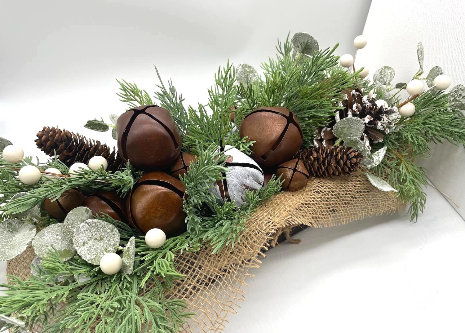 winter centerpiece with metal bells in brown and white, variety of pinecones and winter greenery in long wooden planter in espresso color decorated with burlap