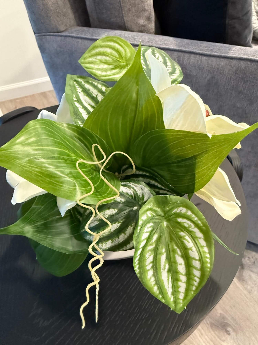 side view of small realistic looking white orchid arrangement with greenery in white ceramic pot vase of geometric shape set on coffee table
