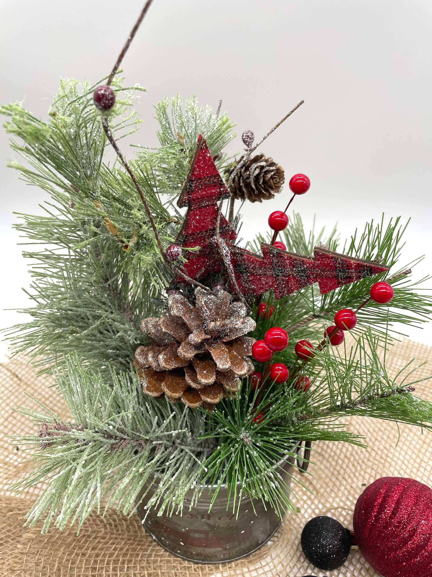 small Christmas arrangement with variety of winter greenery, red ornaments, pinecones in a metal tin with red truck