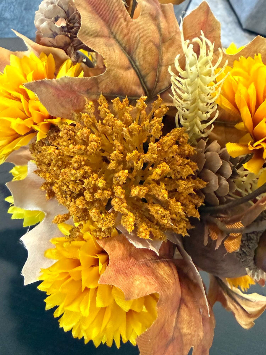 close up for lush elegant handmade small fall arrangement with yellow and gold flowers, brown leaves, poppy heads, acorns, pinecones in white ceramic pumpkin shaped vase
