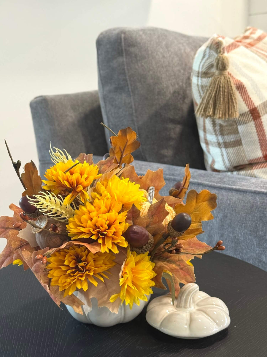 lush elegant handmade small fall arrangement with yellow and gold flowers, brown leaves, poppy heads, acorns, pinecones in white ceramic pumpkin shaped vase styled on coffee table