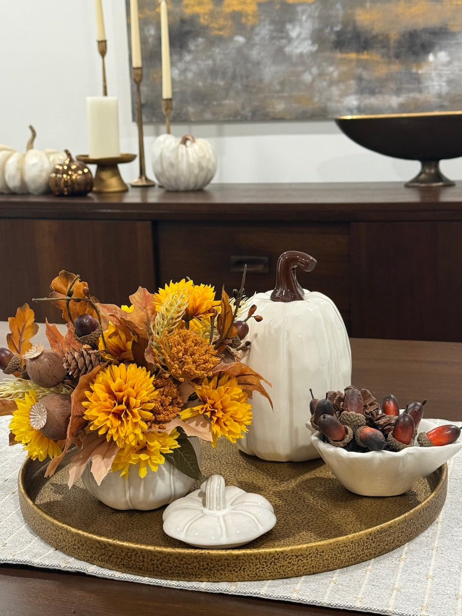 small fall arrangement with yellow flowers, brown and beige fall leaves, poppy heads, acorn stems all in white ceramic pumpkin shape vase styled on tray on dining table