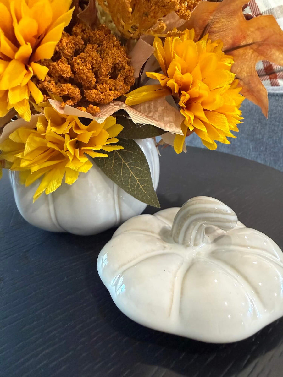 close up for vase of lush elegant handmade small fall arrangement with yellow and gold flowers, brown leaves, poppy heads, acorns, pinecones in white ceramic pumpkin shaped vase styled on coffee table