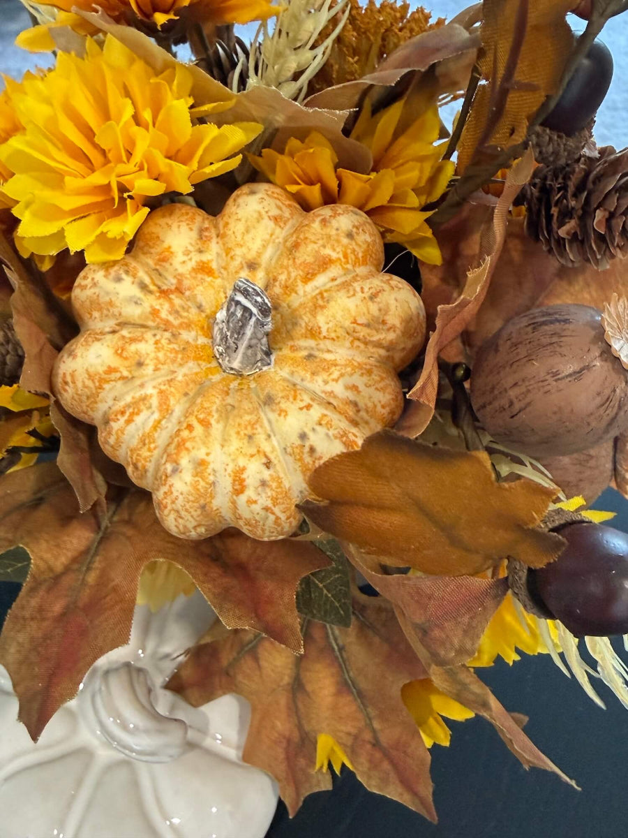 close up lush elegant handmade small fall arrangement with yellow and gold flowers, brown leaves, poppy heads, acorns, pinecones in white ceramic pumpkin shaped vase styled on coffee table