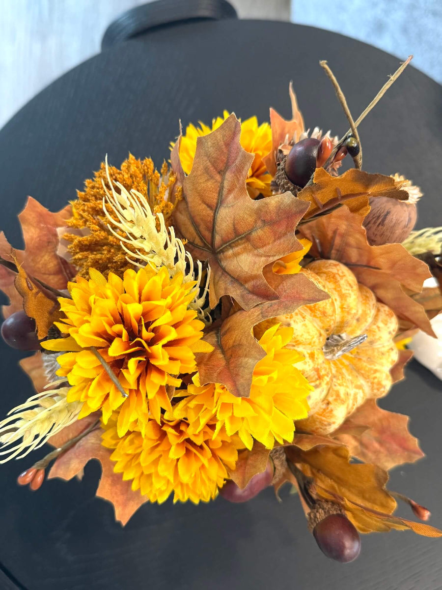 view from top of lush elegant handmade small fall arrangement with yellow and gold flowers, brown leaves, poppy heads, acorns, pinecones in white ceramic pumpkin shaped vase