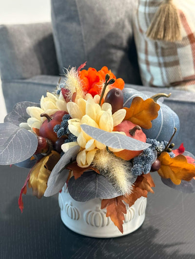 small fall arrangement with blue pumpkin and blue eucalyptus fall flowers in orange and cream, orange/brown little pumpkins wheat acorn in white cearmic vase with pumpkin design set on coffee table